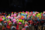 A diverse collection of colorful, small toy figurines placed closely together on sticks. The figurines are bright and varied, featuring neon shades of pink, yellow, green, blue, and orange. The background is dark, which makes the vivid colors of the toys stand out even more.