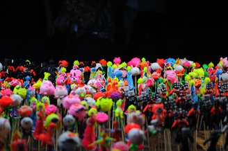 A diverse collection of colorful, small toy figurines placed closely together on sticks. The figurines are bright and varied, featuring neon shades of pink, yellow, green, blue, and orange. The background is dark, which makes the vivid colors of the toys stand out even more.