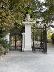 An ornate black wrought iron gate set between white stone pillars opens to a park area with trees. The gate has intricate designs and a stone urn sits atop one of the pillars. The area is surrounded by greenery with a paved path leading up to the gate.