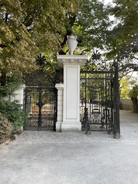 An ornate black wrought iron gate set between white stone pillars opens to a park area with trees. The gate has intricate designs and a stone urn sits atop one of the pillars. The area is surrounded by greenery with a paved path leading up to the gate.