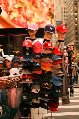A street vendor display with mannequins wearing baseball caps. The caps are primarily branded with the New York Yankees logo in various colors. Behind the display, a blurred, brightly colored billboard is visible, along with some urban architecture. The display includes a mix of hats and clothing items neatly arranged.
