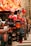 A street vendor display with mannequins wearing baseball caps. The caps are primarily branded with the New York Yankees logo in various colors. Behind the display, a blurred, brightly colored billboard is visible, along with some urban architecture. The display includes a mix of hats and clothing items neatly arranged.