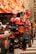 A street vendor display with mannequins wearing baseball caps. The caps are primarily branded with the New York Yankees logo in various colors. Behind the display, a blurred, brightly colored billboard is visible, along with some urban architecture. The display includes a mix of hats and clothing items neatly arranged.
