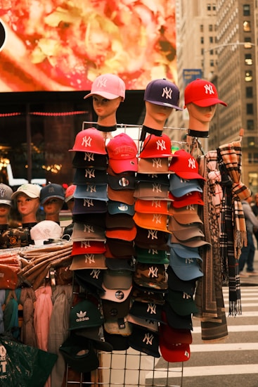 A street vendor display with mannequins wearing baseball caps. The caps are primarily branded with the New York Yankees logo in various colors. Behind the display, a blurred, brightly colored billboard is visible, along with some urban architecture. The display includes a mix of hats and clothing items neatly arranged.