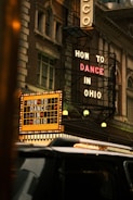A classic theater facade with vintage lighting displays two prominent signs, advertising 'How to Dance in Ohio'. The building’s architecture has an old-fashioned charm, featuring intricate designs and large windows. A black vehicle is visible in the foreground, partially obscuring part of the theater's entrance.
