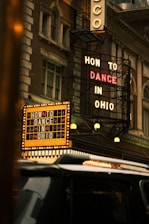 A classic theater facade with vintage lighting displays two prominent signs, advertising 'How to Dance in Ohio'. The building’s architecture has an old-fashioned charm, featuring intricate designs and large windows. A black vehicle is visible in the foreground, partially obscuring part of the theater's entrance.