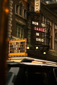 A classic theater facade with vintage lighting displays two prominent signs, advertising 'How to Dance in Ohio'. The building’s architecture has an old-fashioned charm, featuring intricate designs and large windows. A black vehicle is visible in the foreground, partially obscuring part of the theater's entrance.
