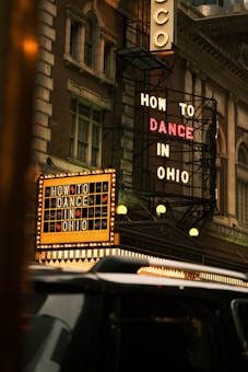 A classic theater facade with vintage lighting displays two prominent signs, advertising 'How to Dance in Ohio'. The building’s architecture has an old-fashioned charm, featuring intricate designs and large windows. A black vehicle is visible in the foreground, partially obscuring part of the theater's entrance.