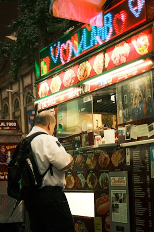 A person wearing a white shirt and a black backpack stands in front of a brightly lit street food truck. The truck displays a variety of food images and colorful LED signs, with words in neon colors. There are multiple food options visible, including wraps and other dishes.