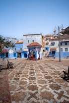 A picturesque plaza characterized by intricate patterned paving stones in a vibrant urban setting. The square is surrounded by buildings painted in varying shades of blue and white, with terracotta roofs. A central structure with a red roof and blue tile accents is a focal point, while potted plants and benches line the area. Two people are seen walking near the structure.