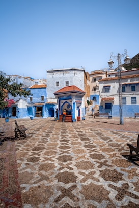 A picturesque plaza characterized by intricate patterned paving stones in a vibrant urban setting. The square is surrounded by buildings painted in varying shades of blue and white, with terracotta roofs. A central structure with a red roof and blue tile accents is a focal point, while potted plants and benches line the area. Two people are seen walking near the structure.