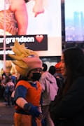 A person dressed in a colorful anime-style costume with spiky hair interacts with someone in a busy public space. A large digital billboard in the background displays a social media post featuring a baby and a heart emoji.