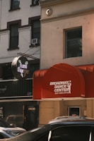 A street view featuring a red awning with the words 'Broadway Dance Center New York City' on it. A neon sign for 'Flash Dancers' is visible. Surrounding buildings have a classic urban architecture with large windows and some visible air conditioning units. Several parked cars can be seen at the bottom, partially in view.