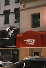 A street view featuring a red awning with the words 'Broadway Dance Center New York City' on it. A neon sign for 'Flash Dancers' is visible. Surrounding buildings have a classic urban architecture with large windows and some visible air conditioning units. Several parked cars can be seen at the bottom, partially in view.