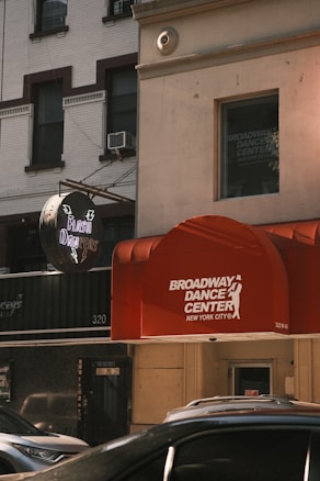 A street view featuring a red awning with the words 'Broadway Dance Center New York City' on it. A neon sign for 'Flash Dancers' is visible. Surrounding buildings have a classic urban architecture with large windows and some visible air conditioning units. Several parked cars can be seen at the bottom, partially in view.