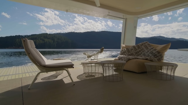 A modern outdoor seating area overlooking a scenic lake and forested hills in the background. The space features comfortable lounge chairs and ottomans with soft cushions arranged on a patio, covered by a roof. Wicker tables are placed near the seating, and a large, cozy blanket is draped over one of the seats. The calm water of the lake reflects the scattered clouds in a bright sky.
