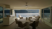 A modern, minimalistic interior with large windows overlooks a serene body of water. The interior features neutral-toned furniture including a round table, chairs, and a cozy seating area with throw pillows. The room is well-lit, emphasizing the spaciousness and inviting atmosphere. The view outside the windows showcases a peaceful lake and distant green hills.