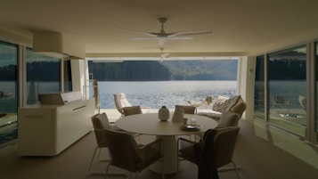 A modern, minimalistic interior with large windows overlooks a serene body of water. The interior features neutral-toned furniture including a round table, chairs, and a cozy seating area with throw pillows. The room is well-lit, emphasizing the spaciousness and inviting atmosphere. The view outside the windows showcases a peaceful lake and distant green hills.