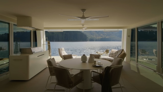 A modern, minimalistic interior with large windows overlooks a serene body of water. The interior features neutral-toned furniture including a round table, chairs, and a cozy seating area with throw pillows. The room is well-lit, emphasizing the spaciousness and inviting atmosphere. The view outside the windows showcases a peaceful lake and distant green hills.