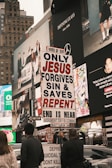 A city street scene with several people holding a religious protest sign that reads 'ONLY JESUS FORGIVES SIN & SAVES REPENT END IS NEAR JUDGMENT DAY IS COMING MATTHEW 24'. The background features large advertisements on a building, depicting fashion and lifestyle imagery. The environment appears urban and busy.