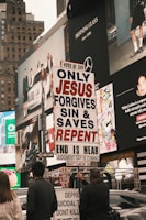 A city street scene with several people holding a religious protest sign that reads 'ONLY JESUS FORGIVES SIN & SAVES REPENT END IS NEAR JUDGMENT DAY IS COMING MATTHEW 24'. The background features large advertisements on a building, depicting fashion and lifestyle imagery. The environment appears urban and busy.