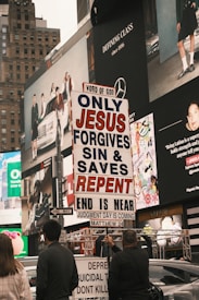 A city street scene with several people holding a religious protest sign that reads 'ONLY JESUS FORGIVES SIN & SAVES REPENT END IS NEAR JUDGMENT DAY IS COMING MATTHEW 24'. The background features large advertisements on a building, depicting fashion and lifestyle imagery. The environment appears urban and busy.