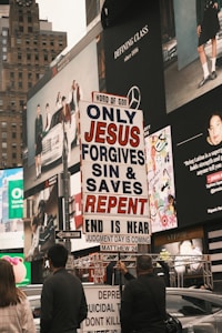 A city street scene with several people holding a religious protest sign that reads 'ONLY JESUS FORGIVES SIN & SAVES REPENT END IS NEAR JUDGMENT DAY IS COMING MATTHEW 24'. The background features large advertisements on a building, depicting fashion and lifestyle imagery. The environment appears urban and busy.