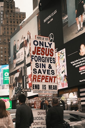 A city street scene with several people holding a religious protest sign that reads 'ONLY JESUS FORGIVES SIN & SAVES REPENT END IS NEAR JUDGMENT DAY IS COMING MATTHEW 24'. The background features large advertisements on a building, depicting fashion and lifestyle imagery. The environment appears urban and busy.