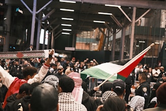 A gathered group of people, some wearing patterned scarves, are participating in a demonstration. A person is holding a flag with distinct colors, and another is holding up a patterned scarf. Police officers are present, monitoring the situation in a city setting under an industrial-looking overhead structure. The crowd appears engaged and focused on the activity.