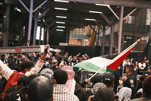 A gathered group of people, some wearing patterned scarves, are participating in a demonstration. A person is holding a flag with distinct colors, and another is holding up a patterned scarf. Police officers are present, monitoring the situation in a city setting under an industrial-looking overhead structure. The crowd appears engaged and focused on the activity.