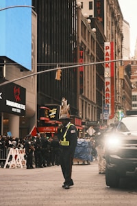 A busy urban street scene with a group of uniformed officers standing in front of various storefronts. A security guard is positioned in the center, wearing a reflective vest marked 'Square'. A large black vehicle with its headlights on is parked on the right. Bright signage and advertisements line the buildings, and there are barriers set up on the sidewalk.