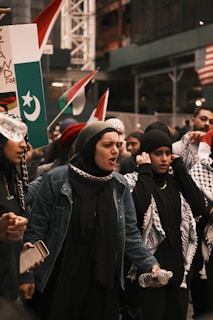 A group of people are participating in a protest or demonstration. They appear to be emotionally charged, with one person holding a sign that displays flags and wearing scarves. The setting seems urban with flags from multiple countries in the background.