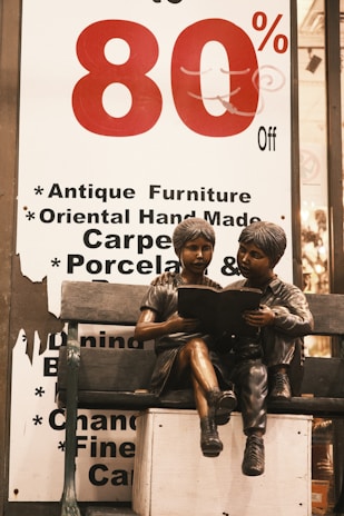A large sale sign with '80% Off' in bold red and black lettering is displayed. Below the sign, a bronze sculpture of two children sitting on a bench is shown, with one child reading a book to the other.