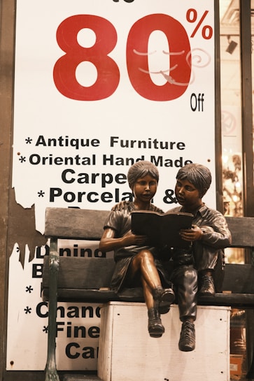 A large sale sign with '80% Off' in bold red and black lettering is displayed. Below the sign, a bronze sculpture of two children sitting on a bench is shown, with one child reading a book to the other.