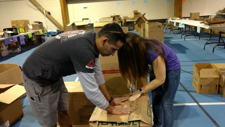 Two people are actively assembling cardboard boxes in a spacious room that appears to be set up for an event or activity. Various open and closed boxes are scattered around, and tables with items and labels can be seen in the background.