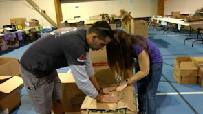 Two people are actively assembling cardboard boxes in a spacious room that appears to be set up for an event or activity. Various open and closed boxes are scattered around, and tables with items and labels can be seen in the background.