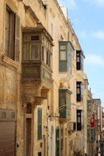 A row of old, stone buildings with traditional Maltese balconies in a narrow street. The buildings are painted in earthy tones with green, red, and black enclosed wooden balconies protruding from the facades. The street is bathed in sunlight, casting shadows and creating a warm, historic atmosphere. There are a few small potted plants and various old doors and windows visible.