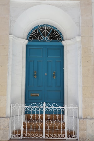 A traditional arched doorway featuring a pair of vibrant blue double doors with ornate golden handles and a mail slot. Above the doors, an intricate wrought iron design is situated in the arch. The doorway is set within a light-colored stone facade, with a small set of steps leading up to it, bordered by a decorative white metal railing.