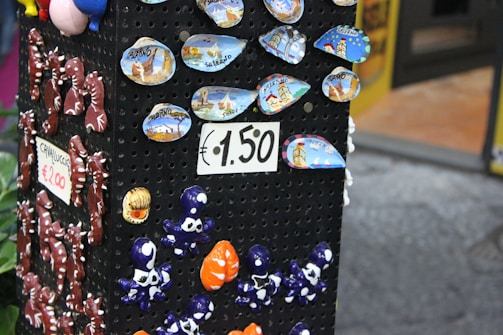 A display stand showcases colorful ceramic souvenirs, including small plates with scenic designs labeled 'Salerno' and various small, whimsical figurines like seahorses and octopuses. Signs indicate prices of €1.50 and €2.00.