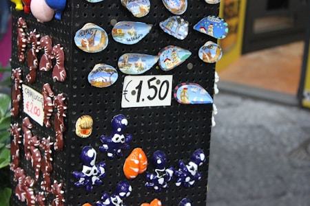 A display stand showcases colorful ceramic souvenirs, including small plates with scenic designs labeled 'Salerno' and various small, whimsical figurines like seahorses and octopuses. Signs indicate prices of €1.50 and €2.00.
