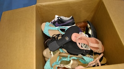 A cardboard box filled with a variety of shoes including sandals, sneakers, and heels in different colors and styles.