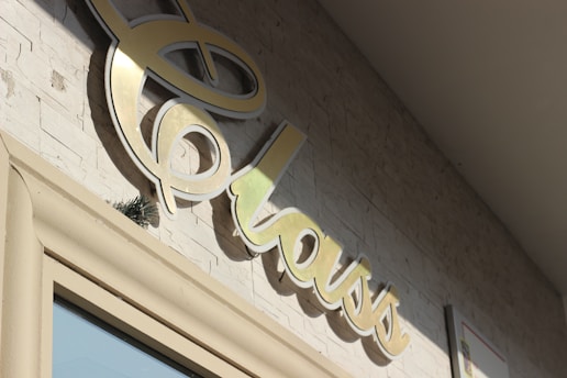 A close-up of a stylized, cursive sign that reads 'class' mounted on a textured stone wall. The sign appears to be made of a shiny, metallic material and is casting a shadow on the wall. The lighting suggests it is a sunny day.