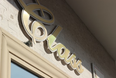 A close-up of a stylized, cursive sign that reads 'class' mounted on a textured stone wall. The sign appears to be made of a shiny, metallic material and is casting a shadow on the wall. The lighting suggests it is a sunny day.