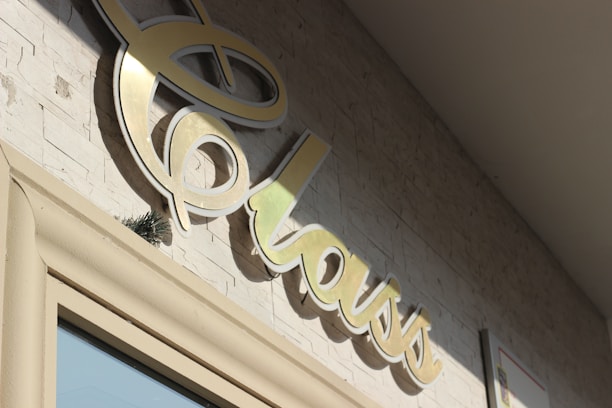 A close-up of a stylized, cursive sign that reads 'class' mounted on a textured stone wall. The sign appears to be made of a shiny, metallic material and is casting a shadow on the wall. The lighting suggests it is a sunny day.