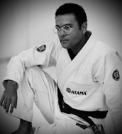 A person is wearing a martial arts gi, adorned with various patches, and is shown in a seated pose. The image features a focused, introspective expression, enhanced by the black belt around the waist. The monochrome style adds a timeless quality.