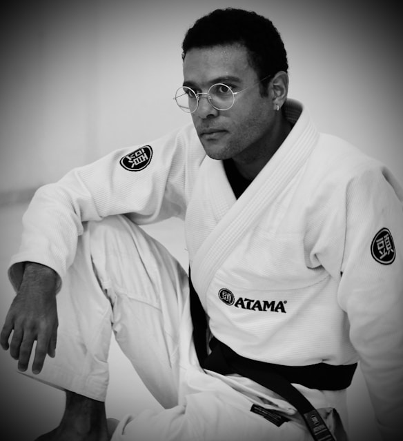 A person is wearing a martial arts gi, adorned with various patches, and is shown in a seated pose. The image features a focused, introspective expression, enhanced by the black belt around the waist. The monochrome style adds a timeless quality.