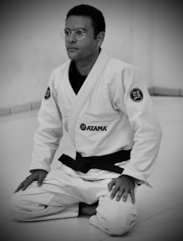 A person wearing a white martial arts gi with a black belt is kneeling on a mat. The gi has patches and the person is wearing round glasses, conveying focus and concentration.