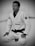 A person wearing a white martial arts gi with a black belt is kneeling on a mat. The gi has patches and the person is wearing round glasses, conveying focus and concentration.