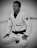 A person wearing a white martial arts gi with a black belt is kneeling on a mat. The gi has patches and the person is wearing round glasses, conveying focus and concentration.