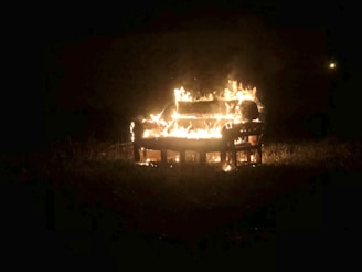 A sofa is engulfed in flames during the night, situated in an open grassy area. The fire illuminates the immediate surroundings, creating a stark contrast against the dark background.